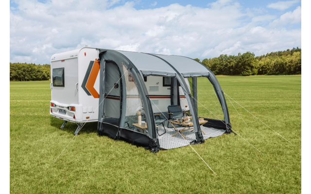 Weißer Wohnwagen mit grauem aufblasbarem Vorzelt; zwei Klappstühle und kleiner Tisch auf einer Unterlage, Wiese, Bäume und wolkiger Himmel im Hintergrund