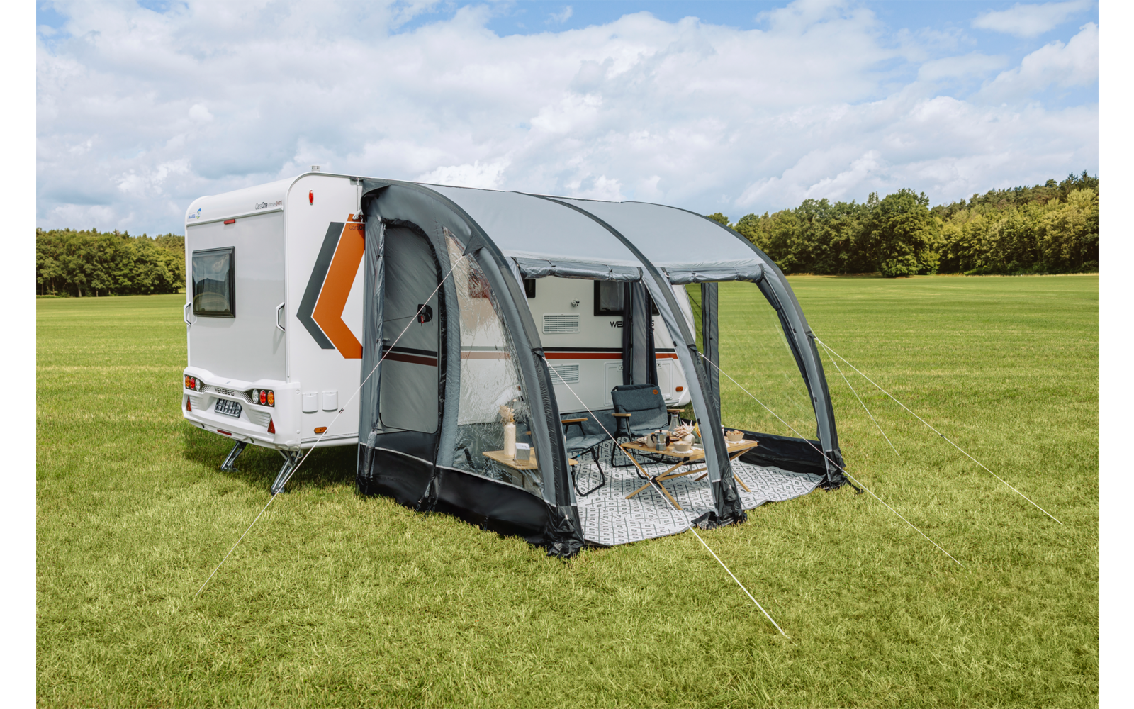 Weißer Wohnwagen mit grauem aufblasbarem Vorzelt; zwei Klappstühle und kleiner Tisch auf einer Unterlage, Wiese, Bäume und wolkiger Himmel im Hintergrund