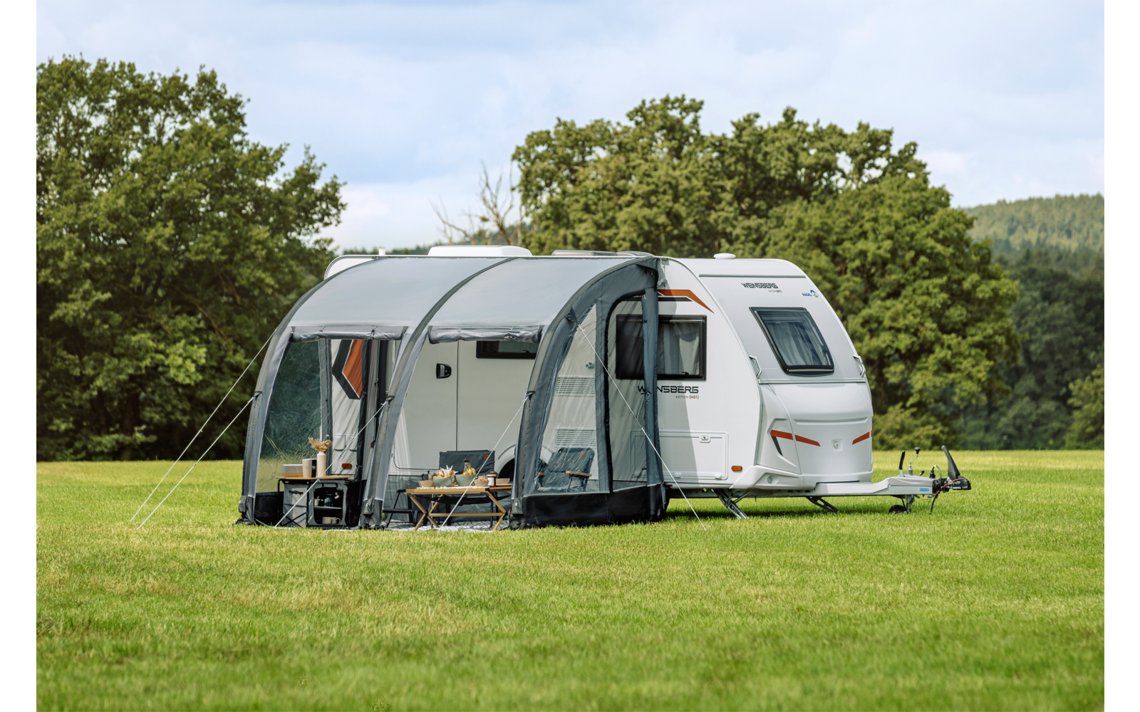Caravan bianco con tendalino scuro, due sedie da campeggio e un tavolino su prato verde, alberi sullo sfondo, cielo nuvoloso