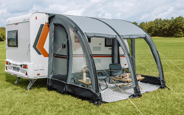 Witte caravan met grijs opblaasbaar voortent; twee klapstoelen en kleine tafel op een mat, grasveld, bomen en bewolkte lucht op de achtergrond