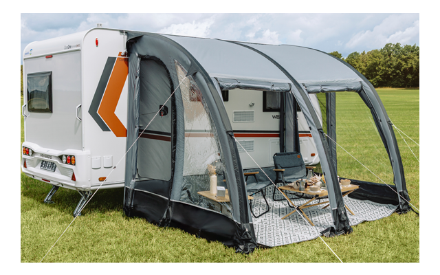 Roulotte bianca con veranda gonfiabile grigia; due sedie pieghevoli e piccolo tavolo su un tappeto, prato, alberi e cielo nuvoloso sullo sfondo