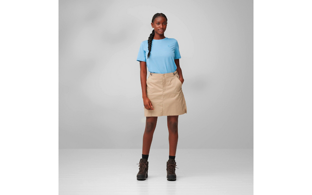Full-length photo of a person standing against a gray background, wearing a blue t-shirt, beige skirt, brown hiking boots, black socks, hair in a braid; hands in pockets