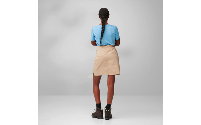 Person seen from behind with long braided hair, light blue T-shirt, beige skirt, black socks and brown boots on a gray background