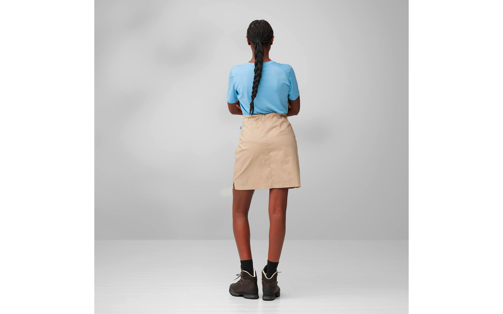 Person seen from behind with long braided hair, light blue T-shirt, beige skirt, black socks and brown boots on a gray background