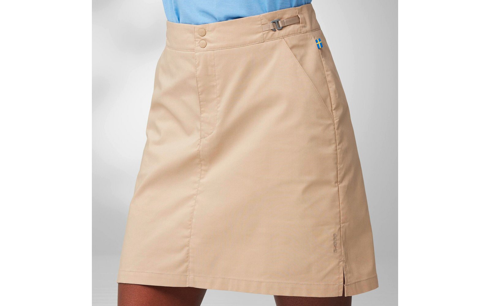 Beige skirt from waist to mid-thigh with side pocket, waist tab showing a small Swedish flag tag, two front snap buttons, light blue top visible at the waist, light gray background