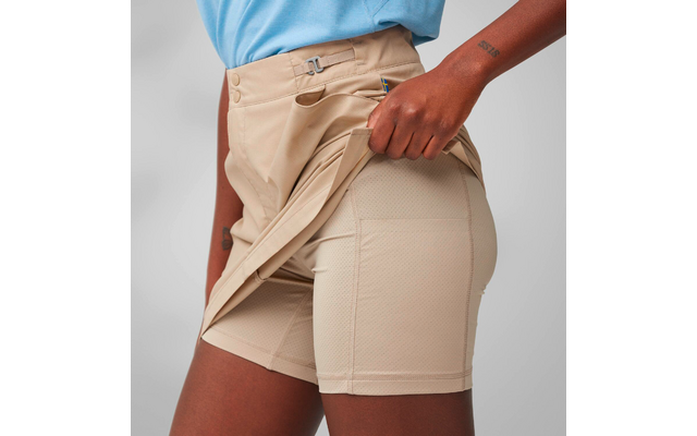 Close-up from waist to mid-thigh of a person. Right hand pulls the beige outer shorts aside, revealing beige fitted inner shorts. Light blue top, metal clasp at the waistband, small blue-yellow label, small tattoo on the forearm