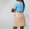 Person seen from back, arms crossed, light blue short-sleeve T-shirt tucked into beige skirt above the knees, long braided hair, gray background