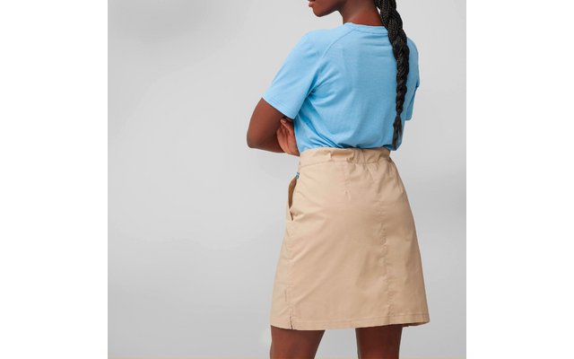 Person seen from back, arms crossed, light blue short-sleeve T-shirt tucked into beige skirt above the knees, long braided hair, gray background