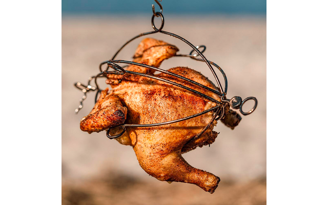 Hele kip met gebruinde huid in een metalen houder, hangend voor een onscherpe strandachtergrond