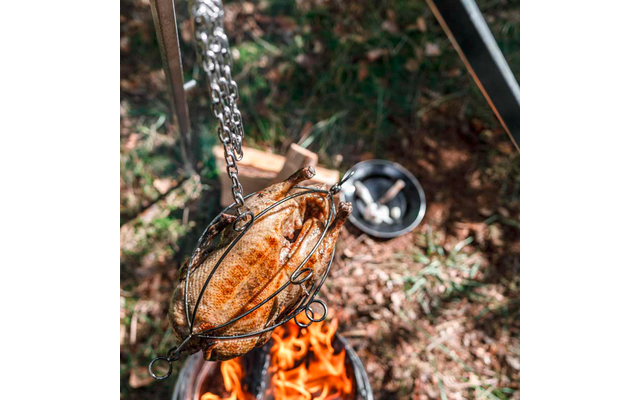 Hele braadkip in een metalen draaispitkorf, aan kettingen boven een open vuur hangend; op de achtergrond een pan met bestek op het gras