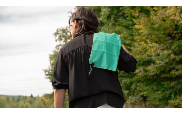 Rückenansicht einer Person in schwarzem Hemd mit einem grünen Handtuch über der Schulter, im Freien vor Bäumen und Himmel