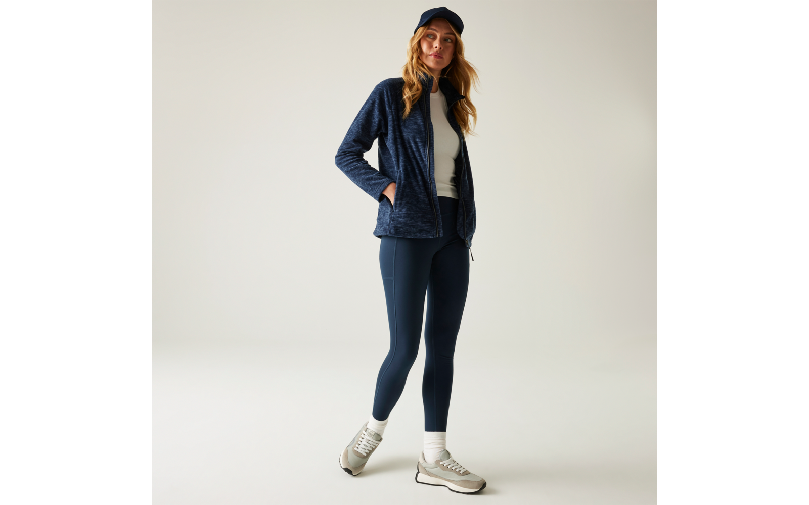 Person in dunkelblauer Fleecejacke, weißem Oberteil, dunkelblauen Leggings, Baseballcap und Turnschuhen, Hände in den Jackentaschen, steht vor hellem Hintergrund