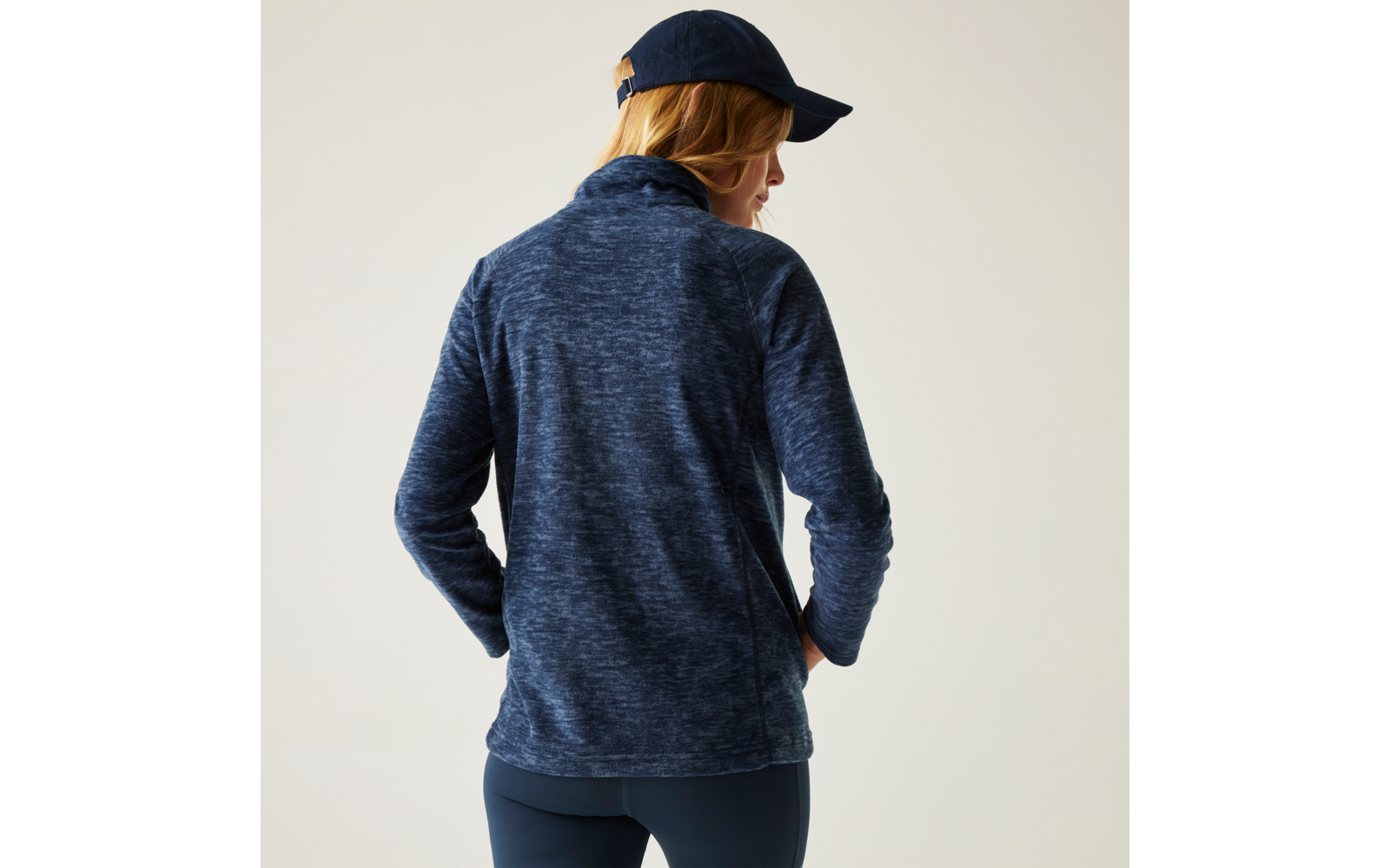 Person von hinten, trägt dunkelblaue Fleecejacke, dunkelblaue Leggings und dunkelblaue Kappe, Hände in den Taschen, heller einfarbiger Hintergrund