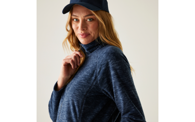 Frau mit blonden Haaren, trägt dunkelblaue Kappe und dunkelblaue Jacke mit Reißverschluss, schaut leicht zur Seite