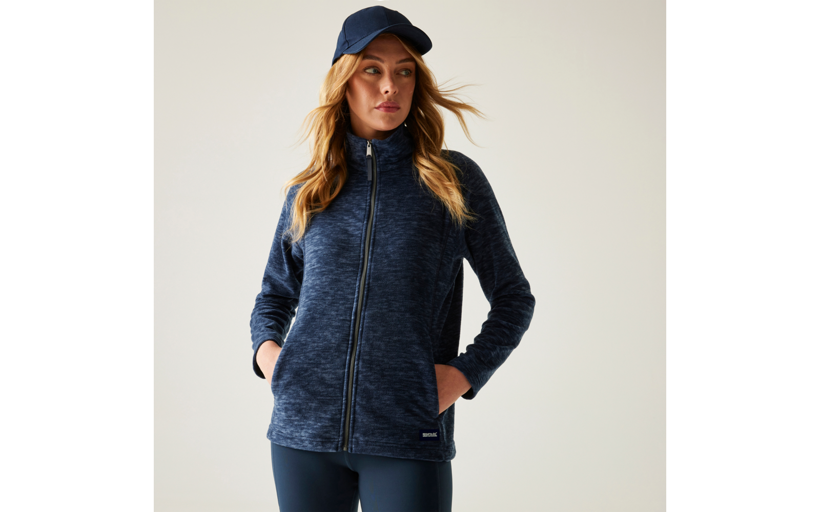 Frau in dunkelblauer Zip‑Up-Fleecejacke und dunkelblauer Baseballkappe, Hände in den Taschen, schaut leicht zur Seite vor hellem Hintergrund
