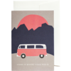 Karte vor weißem Umschlag; auf der Vorderseite ein roter Kreis hinter dunklen Bergsilhouetten, ein rosa Kleinbus mit weißem Dach in Seitenansicht und unten der Schriftzug 'HOME IS WHERE YOUR BUS IS'