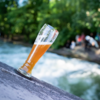 Ein halb volles Bierglas steht schräg auf einem Stein mit einem fließenden Fluss im Hintergrund