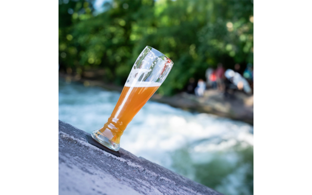 Ein halb volles Bierglas steht schräg auf einem Stein mit einem fließenden Fluss im Hintergrund