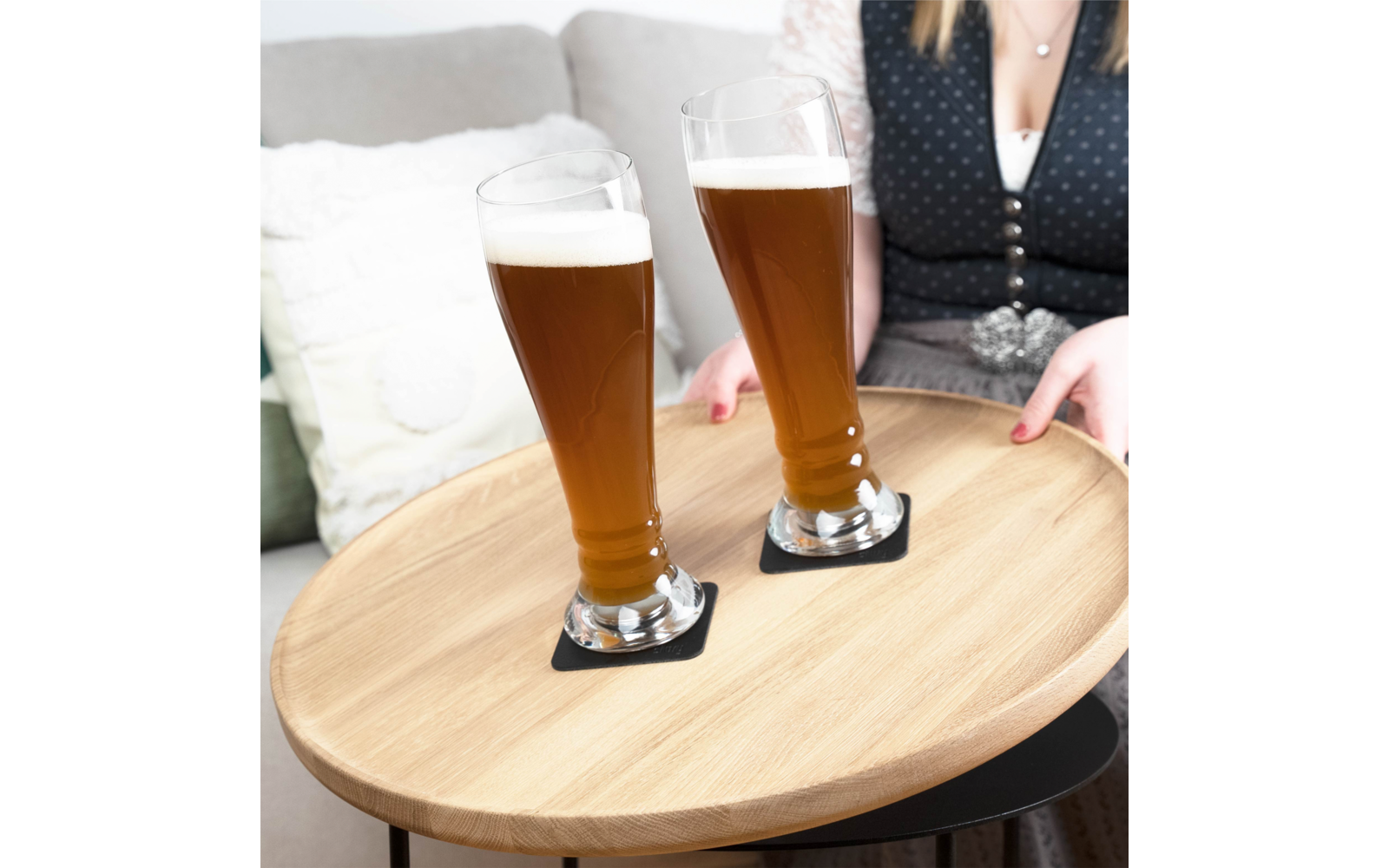 Zwei gefüllte Biergläser auf einem runden Holztablett, weibliche Person im Dirndl im Hintergrund
