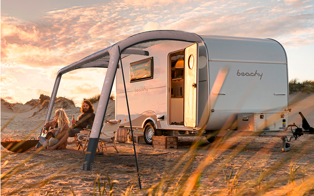 Ein Wohnwagen steht auf sandigem Boden mit ausklappbarem Vorzelt, umgeben von Gras und einem bewölkten Himmel im Hintergrund