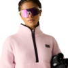 Person mit geflochtenem Haar, rosa Fleecejacke mit halbem Reißverschluss, verspiegelte violette Sonnenbrille, hält ein schwarzes Objekt mit sichtbarem weißem Etikett