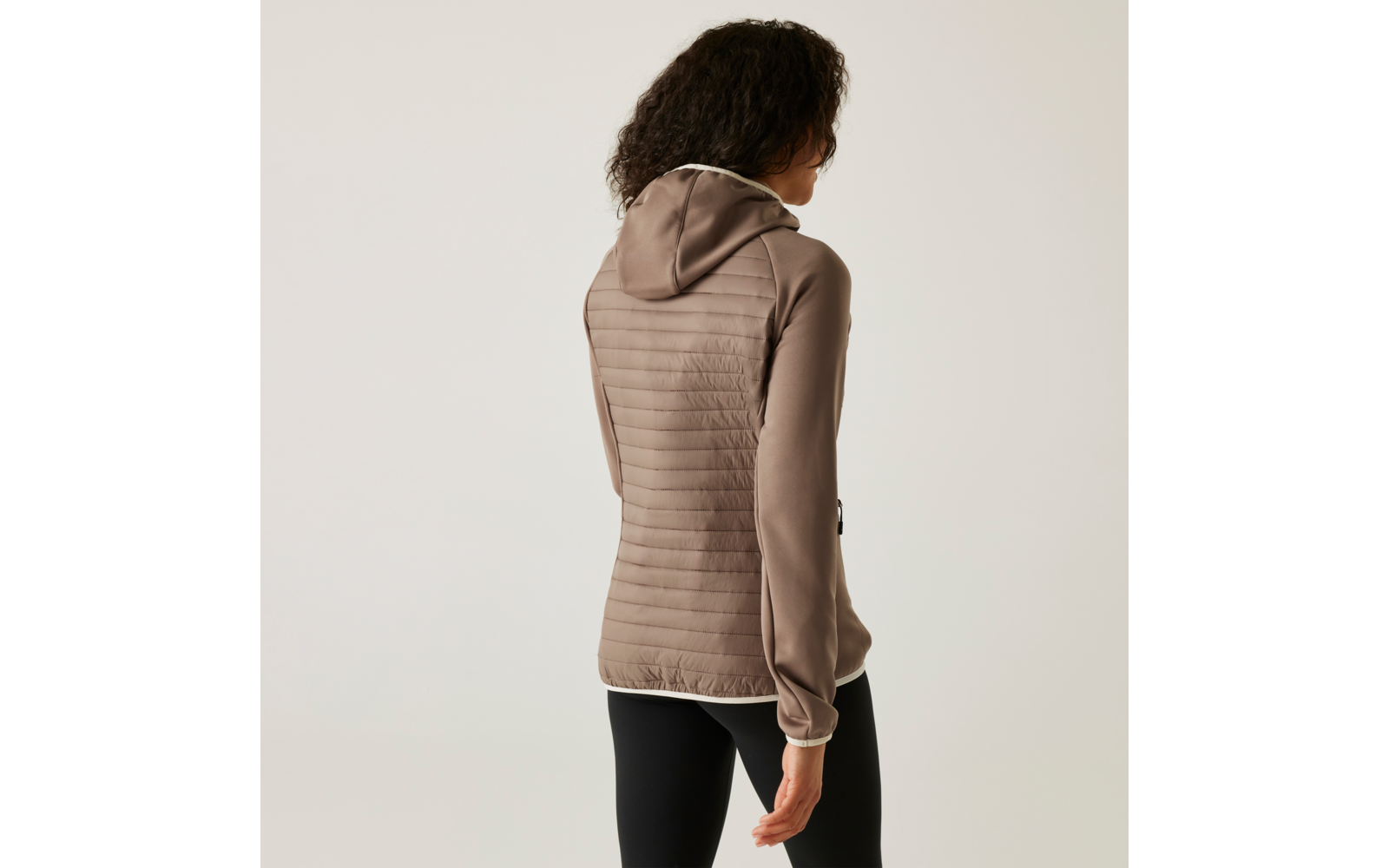 Femme aux cheveux bouclés portant un sweat à capuche beige et un pantalon noir, photographiée de dos