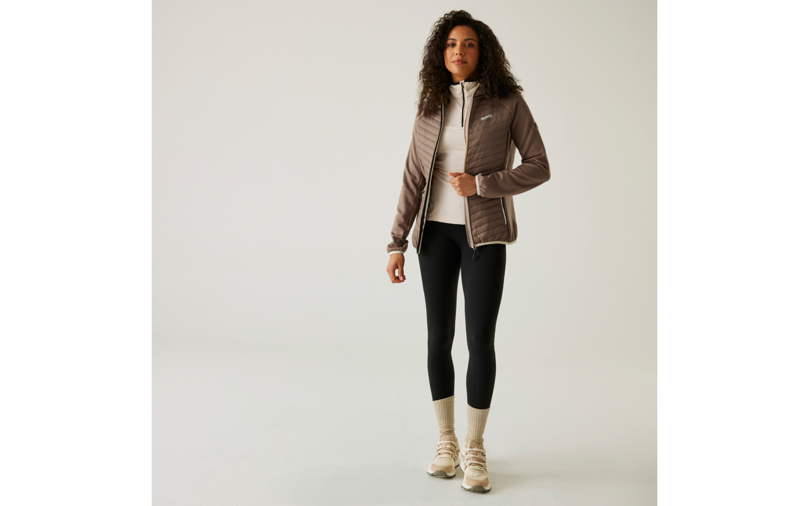Femme portant un blazer marron clair, un pull blanc, un pantalon noir et des bottes claires sur un fond clair