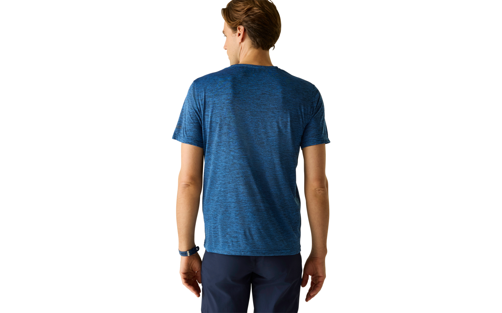 Person von hinten, trägt blaues T‑Shirt mit kurzen Ärmeln, dunkle Hose, Armbanduhr am linken Handgelenk, heller einfarbiger Hintergrund