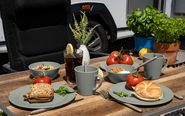 Holztisch mit zwei gedeckten Plätzen: graue Teller mit Brötchen, Schalen mit Salat und Müsli, graue Tassen mit Löffeln, Glas mit dunklem Getränk und Zitronenscheibe, frische Tomaten, Kräuter in Töpfen, schwarzer Stuhl und weißer Wohnwagen im Hintergr