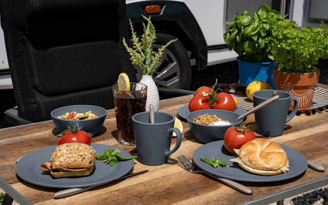 Holztisch vor einem Wohnwagen mit Tellern, Brötchen, Tassen, Schalen, Tomaten und Kräutertöpfen