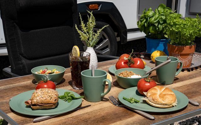 Holztisch im Freien mit zwei grünen Tellern jeweils mit einem Brötchen, zwei grünen Tassen mit Löffeln, ein Glas mit dunkler Flüssigkeit und Zitronenscheibe, Schalen mit Kirschtomaten und Müsli, Kräuter in Töpfen, ein Stuhl und ein Fahrzeug im Hinter