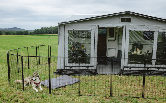 Weißes Vorzelt an einem Wohnwagen mit Fenstern auf einer Wiese; schwarzer Metallzaun umschließt einen Bereich; Hund mit rotem Geschirr liegt auf dem Gras außerhalb des Zauns; bewölkter Himmel und Hügel im Hintergrund
