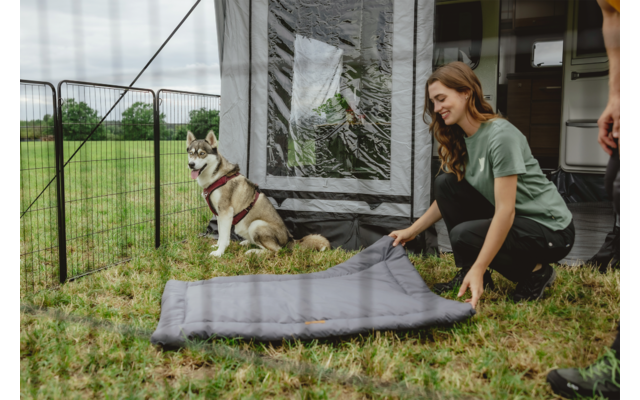 Frau kniet und legt eine zusammengefaltete Isomatte auf Gras vor einem Wohnwagenvorzelt; ein Husky mit Geschirr sitzt neben einem Metallzaun; rechts sind Beine einer weiteren Person sichtbar