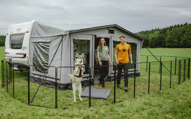 Wohnmobil mit angebautem Vorzelt auf einer Wiese, davor eingezäunter Bereich mit hellem Hund, zwei Personen stehen am Vorzelteingang, im Hintergrund Wald und bewölkter Himmel