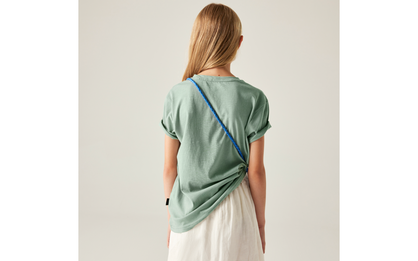 Person von hinten, lange blonde Haare, grünes kurzärmeliges Oberteil, weißer Rock, blauer Riemen diagonal über dem Rücken, heller einfarbiger Hintergrund