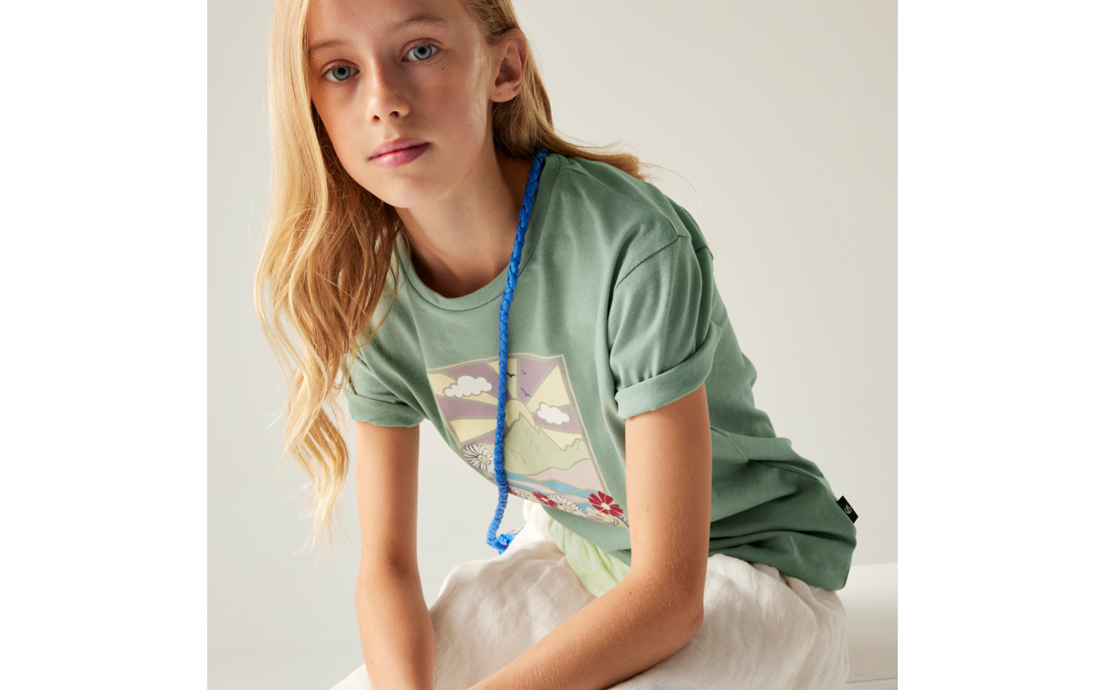 Person mit langen blonden Haaren sitzt, trägt grünes T‑Shirt mit rechteckigem Landschaftsprint und blaue geflochtene Kordel, weiße Rock- oder Stoffhose