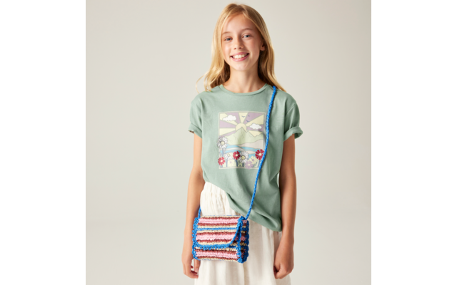 Lächelndes blondes Mädchen, trägt hellgrünes T‑Shirt mit Berg-, Sonne- und Blumenmotiv, weißer Rock, bunte gehäkelte Umhängetasche mit blauem Träger