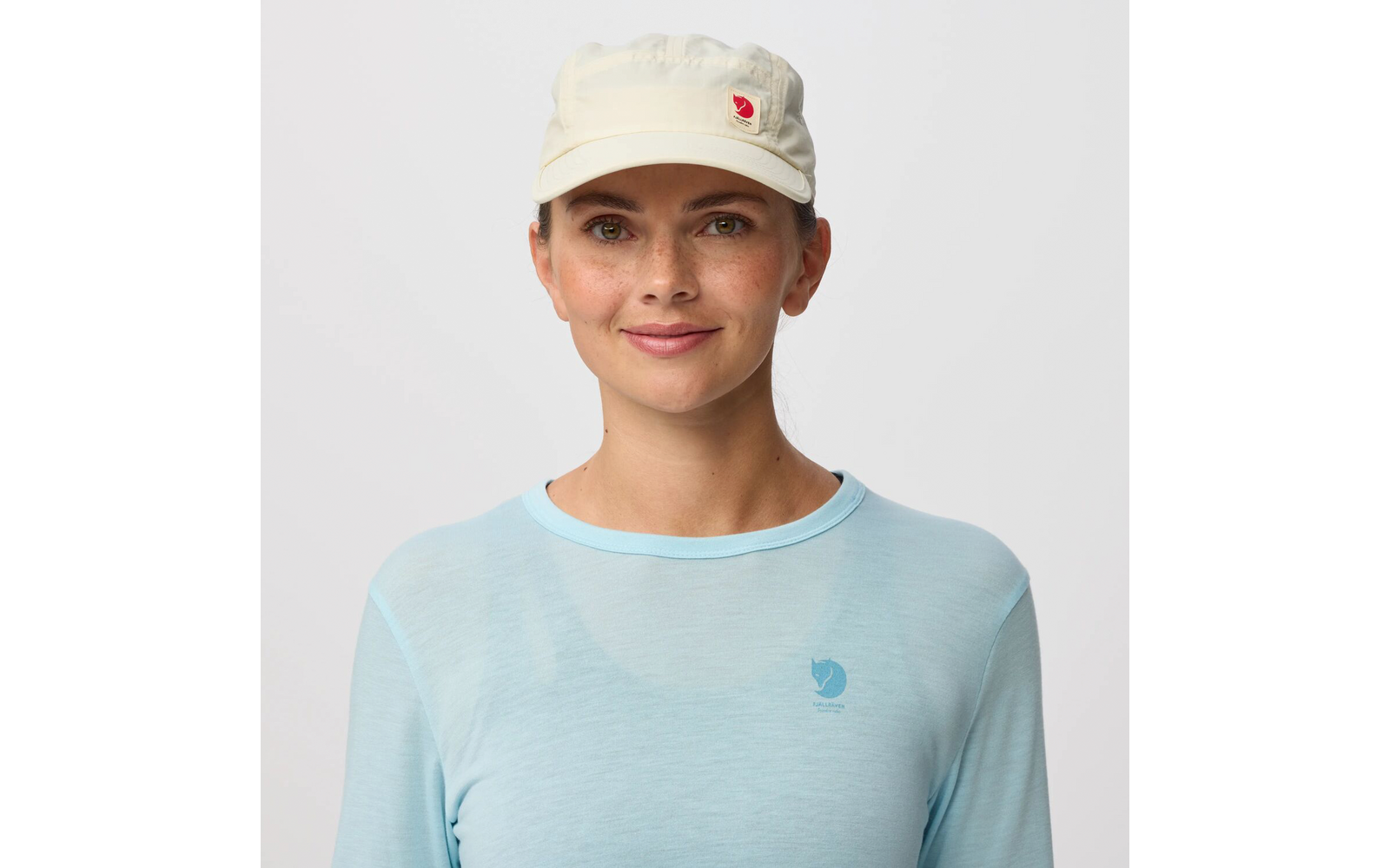 Persona frente a un fondo liso y claro, lleva una gorra color crema con un emblema rojo y una camiseta de manga larga azul claro con un pequeño logo en el pecho