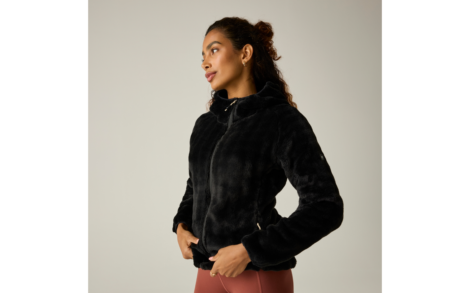 Persona con chaqueta negra y peluda con capucha y cremallera, manos en los bolsillos, mira hacia la izquierda, lleva mallas de color marrón rojizo, fondo liso claro