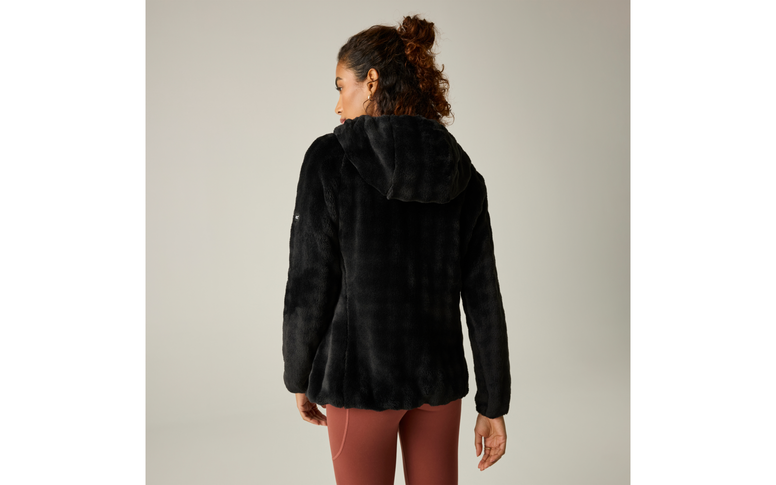 Persona vista de espaldas, lleva una chaqueta con capucha negra de textura aterciopelada y leggings marrón rojizo, pelo recogido en un moño suelto, fondo liso y claro