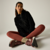 Persona sentada con las piernas cruzadas sobre fondo claro, viste una sudadera negra con cremallera, mallas color óxido, calcetines beige y zapatillas burdeos, pelo recogido en una coleta