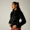 Persona con chaqueta negra y peluda con capucha y cremallera, manos en los bolsillos, mira hacia la izquierda, lleva mallas de color marrón rojizo, fondo liso claro
