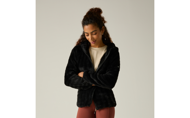 Persona con el pelo recogido lleva una chaqueta polar negra, brazos cruzados, mira hacia abajo, camiseta clara y leggings color óxido sobre fondo gris claro