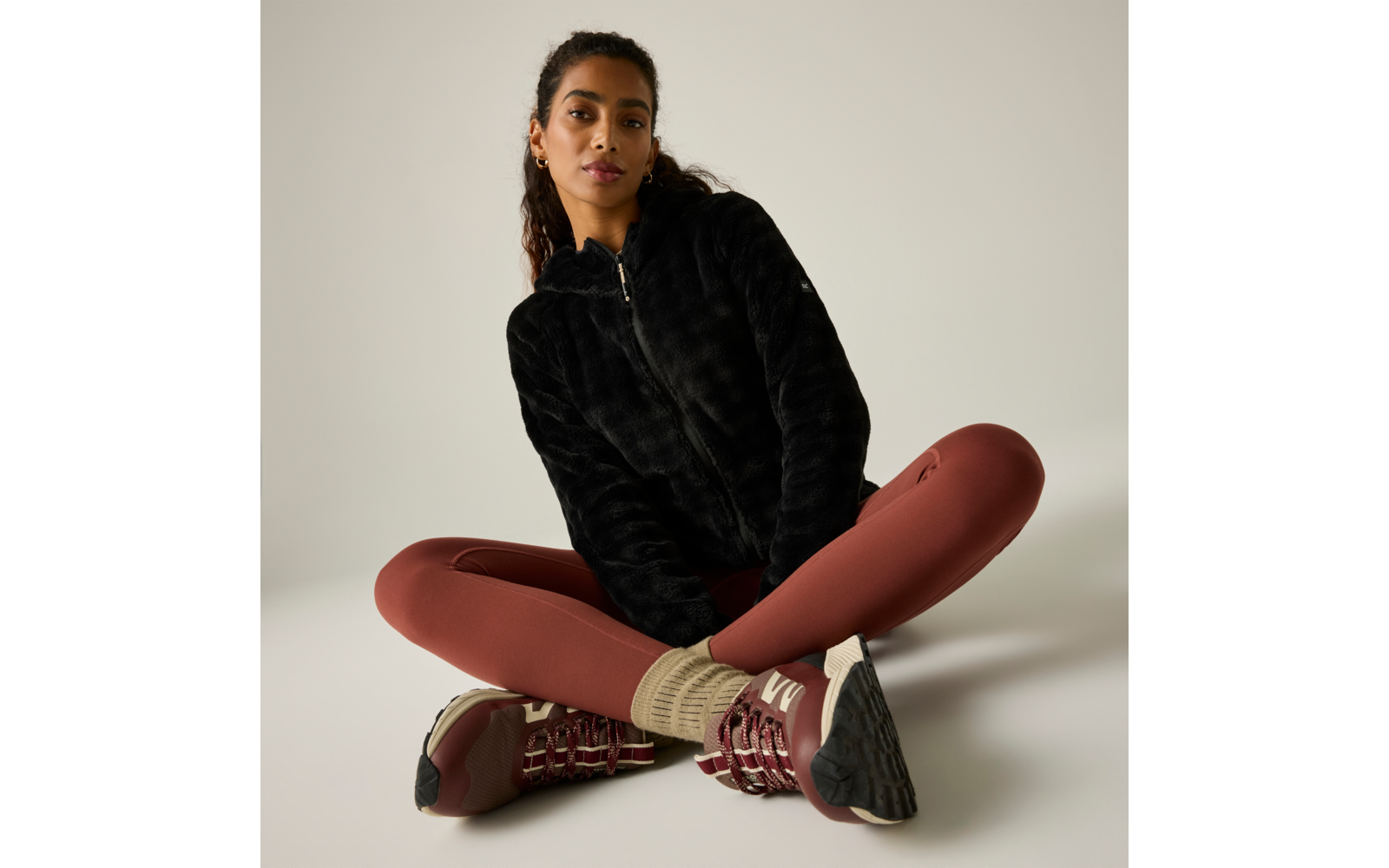 Persona sentada con las piernas cruzadas sobre fondo claro, viste una sudadera negra con cremallera, mallas color óxido, calcetines beige y zapatillas burdeos, pelo recogido en una coleta