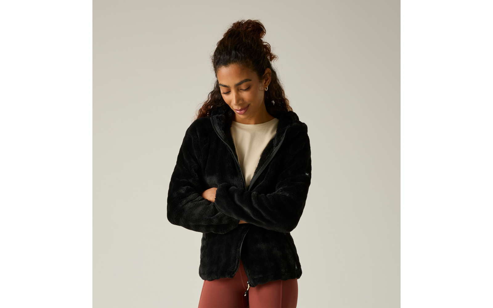 Persona con el pelo recogido lleva una chaqueta polar negra, brazos cruzados, mira hacia abajo, camiseta clara y leggings color óxido sobre fondo gris claro
