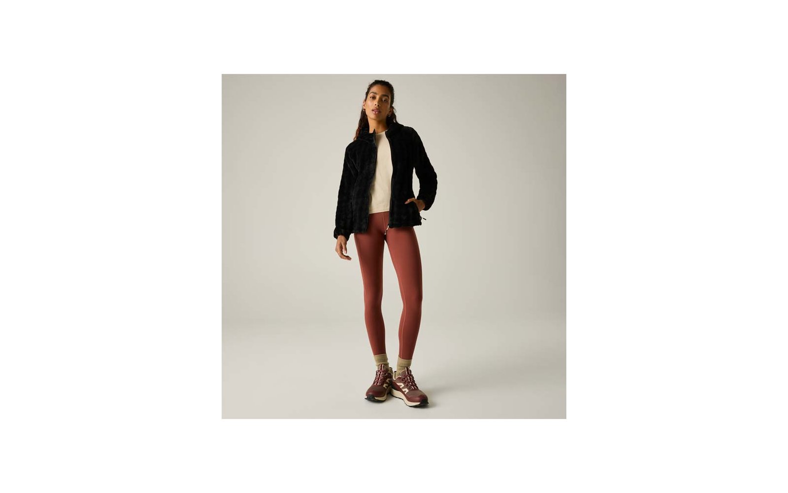 Mujer, cuerpo entero visible, lleva chaqueta negra de pelo sintético, suéter claro, mallas color óxido, zapatillas de senderismo burdeos y calcetines amarillos sobre fondo liso