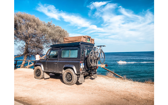 Veicolo fuoristrada nero con tenda da tetto e bicicletta montata sul retro, parcheggiato su terreno sabbioso di una scogliera, mare sullo sfondo, albero a sinistra, corrimano di legno e una persona vicino al corrimano