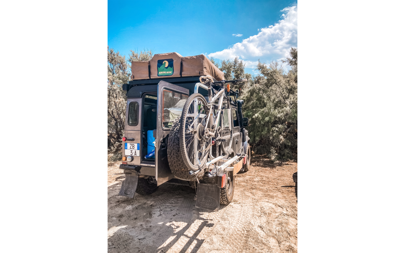 Vista posteriore di un veicolo con tenda da tetto e due biciclette montate sul retro, parcheggiato su terreno sabbioso davanti a cespugli sotto un cielo azzurro con nuvole