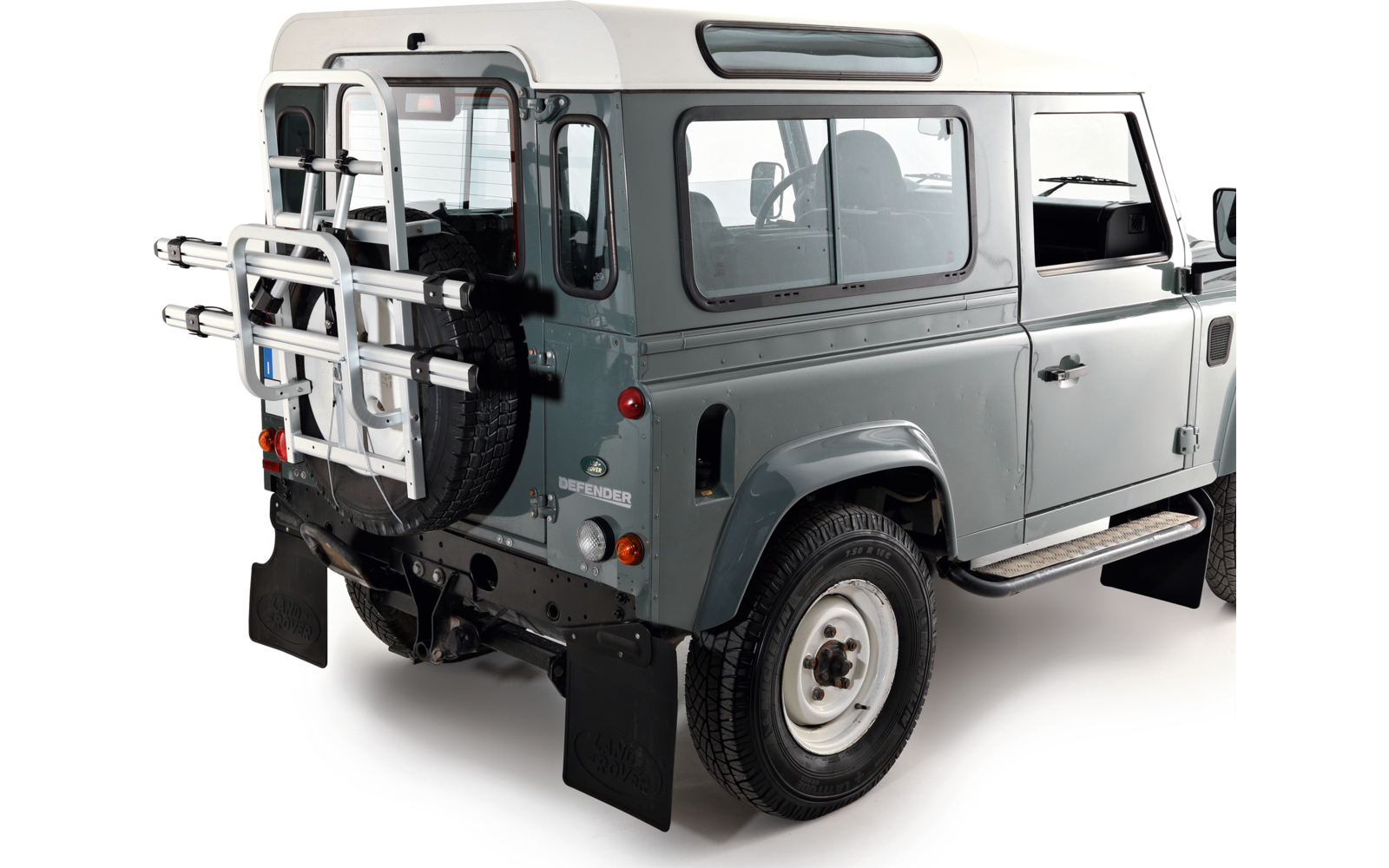 Vista posteriore tre quarti di un veicolo grigio chiaro con tetto bianco, finestrini laterali, ruota di scorta e portapacchi metallico montati sul portellone posteriore, fanali posteriori e pedana