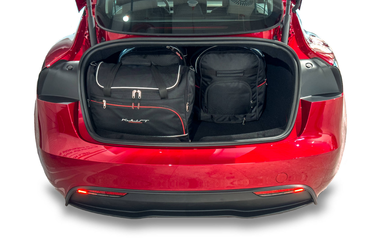 Coffre ouvert d'une voiture rouge avec deux sacs noirs : à gauche un grand sac de voyage avec fermetures éclair blanches et accents rouges, à droite un sac à dos noir